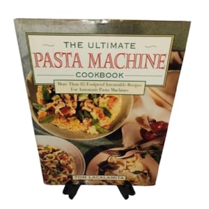 May include: A cookbook titled "The Ultimate Pasta Machine Cookbook" with a green and beige cover. The cover features images of pasta dishes and text that reads "More Than 25 Foolproof Irresistible Recipes For Automatic Pasta Machines" and the author's name, Tom Lacalamita.