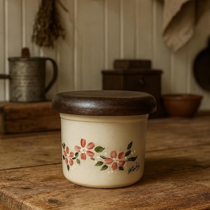May include: A cream-colored ceramic jar with a dark wooden lid. The jar is decorated with pink floral designs and green leaves. The jar is sitting on a wooden table.