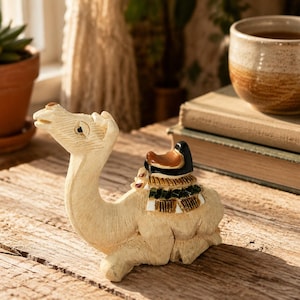 May include: A small, light-colored carved wooden camel figurine in a seated position. The camel features a detailed saddle with black, brown, and white painted accents on its back, showcasing visible wood grain texture and carved facial features.