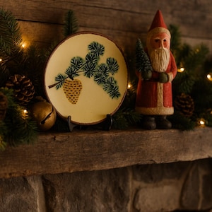 May include: A festive Christmas display featuring a decorative plate with a pine cone and branch design, a Santa Claus figurine holding a small tree, and pine boughs. The plate is on a black stand, and the scene is set on a wooden mantel.
