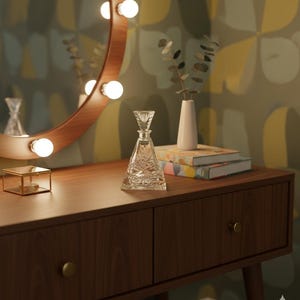 May include: A wooden vanity with a round lighted mirror, a crystal perfume bottle, and a small gold box. The vanity has two drawers with brass knobs. A white vase with greenery and two books are also on the vanity.