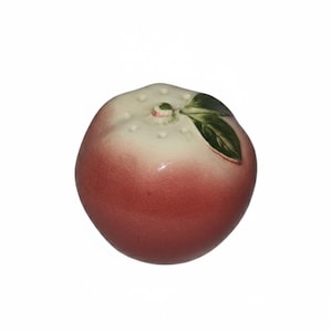 May include: A ceramic peach figurine with a gradient of colors from white to red. The top is white with small, raised dots. A green leaf is attached to the top. The figurine is a decorative item.