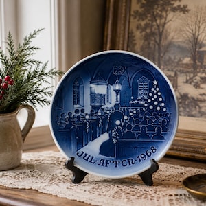May include: A round porcelain decorative plate featuring a deep blue and white relief illustration of a church interior during a Christmas service, with the text JUL AFTEN 1968 printed in a curved font along the bottom edge of the plate.