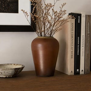 May include: A smooth, matte brown ceramic vase with a tapered base and a wide opening, containing a cluster of dried, light-colored branching flowers. The vase is positioned next to a stack of hardcover books and a small, shallow, textured bowl.