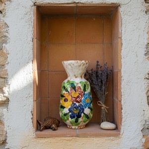 May include: A colorful ceramic vase with a floral design, displayed in a recessed wall niche. The vase features yellow, blue, and red flowers. A small fish figurine, a bundle of lavender, and a smooth stone are also in the niche.
