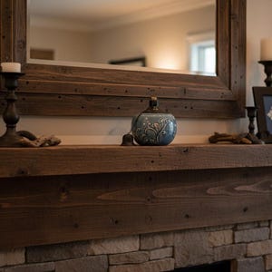 May include: A rustic wooden mantelpiece with a large framed mirror. Decorative items include a blue ceramic vase, framed artwork, and black candle holders with lit candles. The mantel is set above a stone fireplace.
