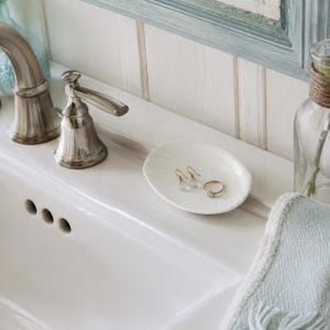 May include: A white ceramic shell-shaped trinket dish holding a pair of light blue teardrop earrings and a gold ring with a small blue stone. The dish is placed on a white porcelain bathroom sink next to a metallic faucet and a light blue towel.