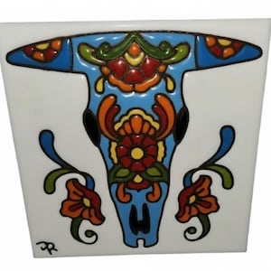 May include: A square, white ceramic tile featuring a stylized bull skull design. The skull is outlined in blue and filled with colorful floral patterns in red, orange, yellow, and green. The design is reminiscent of Day of the Dead art.