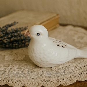 May include: A white ceramic bird figurine with a textured surface, perched on a lace doily. The bird has a small beak and dark eyes. A bundle of dried lavender and a book are in the background.