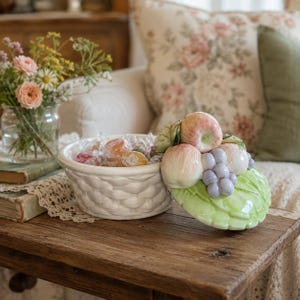 May include: A white ceramic basket-weave bowl filled with individually wrapped candies. The matching lid features a sculpted arrangement of painted ceramic fruit, including apples and grapes, resting on a green leaf-shaped base.
