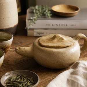 May include: A beige ceramic teapot with a textured, matte finish, a rounded body, a side handle, and a matching lid with a small pointed knob. Two matching ceramic tea cups filled with green tea and a small dish of loose tea leaves are nearby.