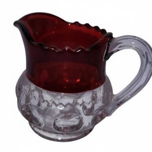 May include: A vintage glass pitcher with a ruby red top and a clear, patterned base. The pitcher has a curved handle and a ruffled rim. The base features a series of embossed circles. This decorative pitcher is ideal for serving beverages.