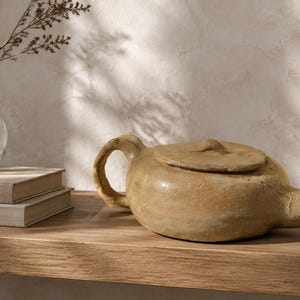 May include: A low-profile, hand-formed tan ceramic teapot with a textured, matte finish, a side handle, and a flat lid with a small central knob. The teapot is positioned next to two stacked beige books and a clear glass vase containing a dried plant stem.