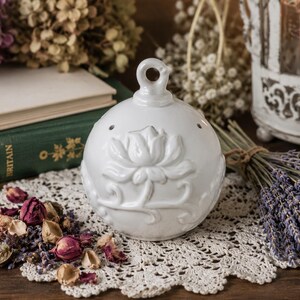 May include: A spherical white ceramic pomander featuring a raised relief floral design of a blooming flower with swirling stems. The object has a small circular loop at the top and several tiny ventilation holes around the upper portion of the sphere.