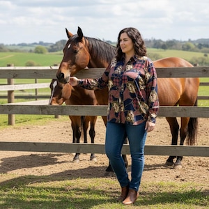 May include: A woman wearing a long-sleeved button-down shirt with a patchwork design in shades of red, blue, and gold, paired with blue jeans and brown boots. Two brown horses stand behind a wooden fence in a rural setting.
