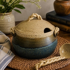 May include: A round ceramic soup tureen with a lid and a matching ladle. The tureen features a speckled beige upper section and a deep blue lower section, with braided ceramic handles and a braided handle on the lid and the ladle.