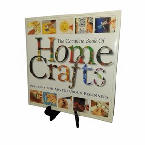 May include: A hardcover book titled "The Complete Book Of Home Crafts" is displayed on a black stand. The title is in large, colorful letters. The book cover features various craft projects and the subtitle "Projects for Adventurous Beginners."