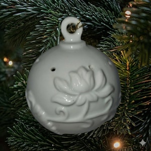 Puede incluir: Adorno navideño de cerámica blanca con diseño floral. El adorno redondo tiene un lazo para colgar y pequeños agujeros. Sobre un fondo de agujas de pino verdes.
