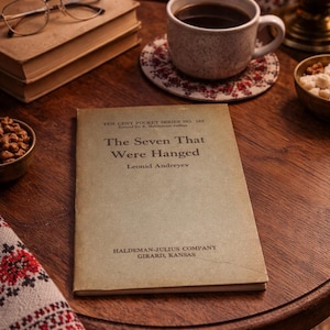 May include: A still life featuring a vintage book titled "The Seven That Were Hanged" by Leonid Andreyev. A cup of coffee sits on a floral coaster, with a small bowl of nuts and a pair of glasses resting on a stack of books.