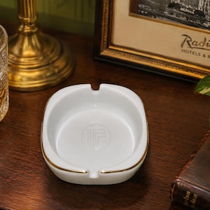 May include: A white ceramic ashtray with a rounded square shape, featuring a gold-trimmed rim and a debossed circular logo in the center. The ashtray has two notches on opposite sides for resting items, with a smooth, glossy finish throughout.