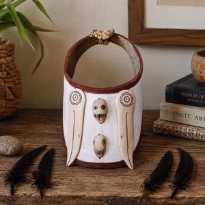 May include: A handmade white and brown decorative basket with a handle. The basket features two stylized bone-colored figures and two eye-shaped designs. Black feathers and a stone are arranged on the wooden surface.