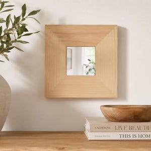 May include: Square wooden framed mirror with a light wood finish. The mirror is mounted on a white wall. A white vase with green leaves and a wooden bowl are on a wooden surface. Books are stacked below the bowl.