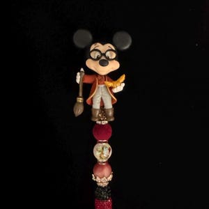 May include: A decorative pen featuring a Mickey Mouse figurine dressed as a wizard. The pen has a red jeweled barrel, a series of decorative beads, and a figure holding a broom and a golden object.