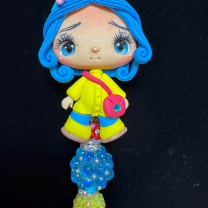 Puede incluir: Figura artesanal de un personaje con cabello azul, que lleva un abrigo amarillo y un bolso rosa. La figura est&aacute; unida a un tallo de cuentas con detalles azules y amarillos. La figura tiene grandes ojos azules y una expresi&oacute;n triste.