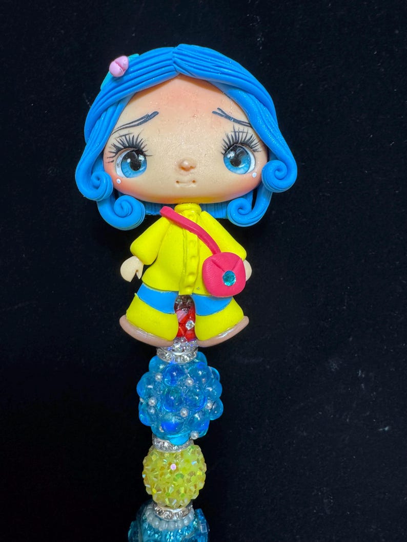 Puede incluir: Figura artesanal de un personaje con cabello azul, que lleva un abrigo amarillo y un bolso rosa. La figura est&aacute; unida a un tallo de cuentas con detalles azules y amarillos. La figura tiene grandes ojos azules y una expresi&oacute;n triste.