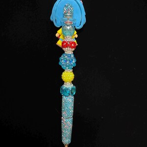 Puede incluir: Un bol&iacute;grafo decorativo con un dise&ntilde;o azul, amarillo y rojo. El bol&iacute;grafo presenta una figura azul en la parte superior, seguida de una serie de cuentas coloridas y un cuerpo brillante de color turquesa. La punta del bol&iacute;grafo es dorada.