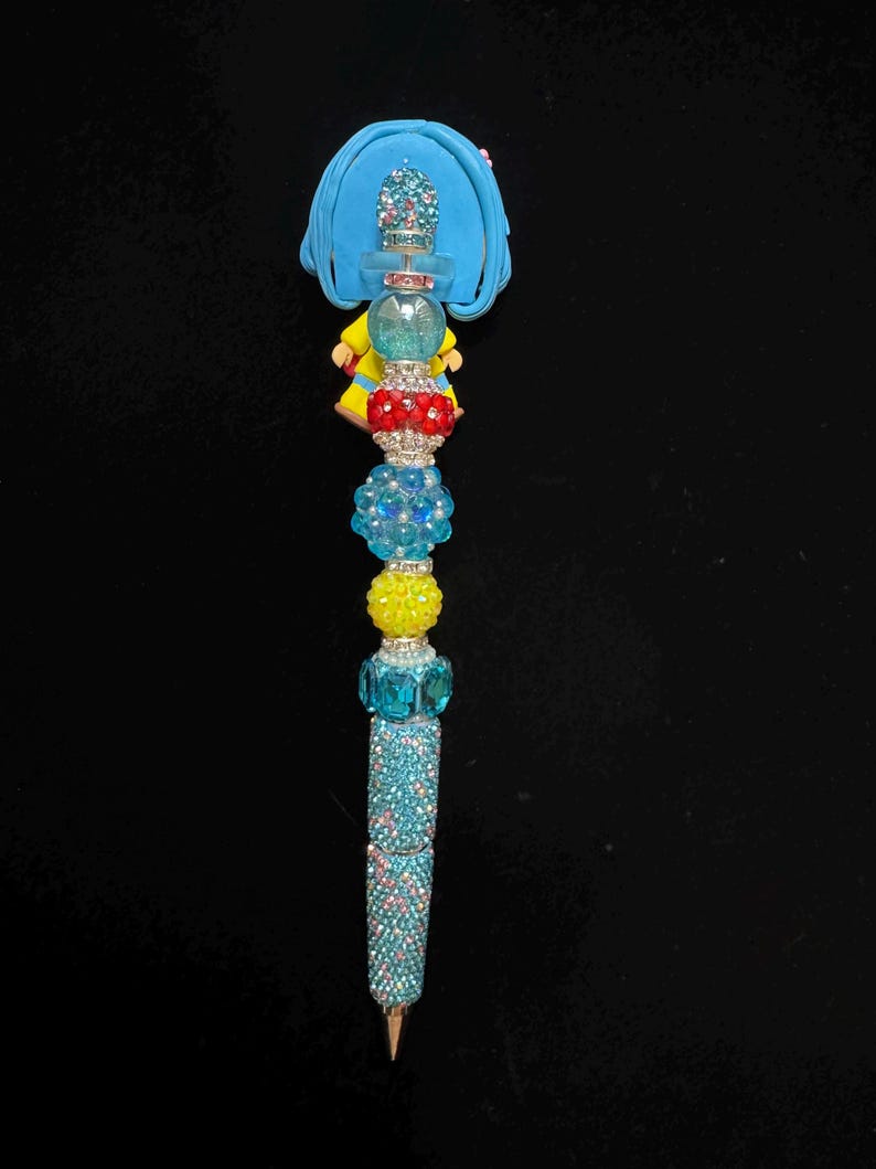 Puede incluir: Un bol&iacute;grafo decorativo con un dise&ntilde;o azul, amarillo y rojo. El bol&iacute;grafo presenta una figura azul en la parte superior, seguida de una serie de cuentas coloridas y un cuerpo brillante de color turquesa. La punta del bol&iacute;grafo es dorada.