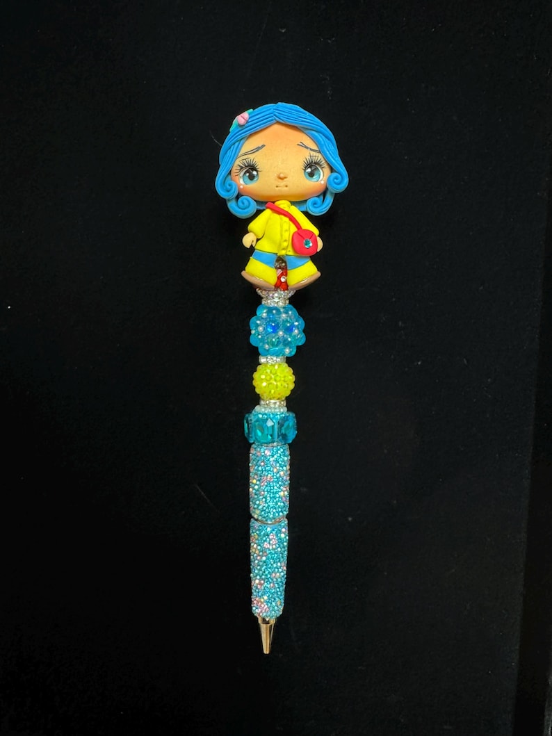 Puede incluir: Bol&iacute;grafo decorativo con un personaje de dibujos animados con pelo azul y abrigo amarillo. El cuerpo del bol&iacute;grafo est&aacute; adornado con brillantes pedrer&iacute;a azul y cuentas azules y amarillas. El bol&iacute;grafo tiene una punta dorada.