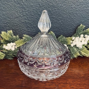 May include: A clear glass candy dish with a removable lid featuring a pointed finial. The bowl and lid have a pressed glass pattern with a band of deep purple color around the center. The glass surface is textured with intricate embossed details.