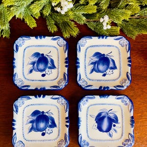 May include: Four square ceramic plates with a white background and blue fruit and leaf design. The plates have a blue border with floral accents. The plates are arranged on a dark wooden surface with greenery in the background.