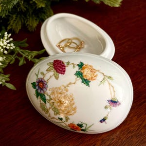 May include: White, oval-shaped trinket box with a floral design in shades of yellow, orange, purple, and red. The lid is open, revealing gold rings inside. The box is on a dark wooden surface.