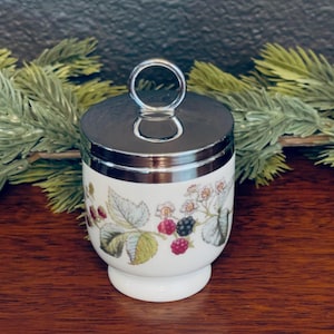 May include: White ceramic egg cup with a silver lid and handle. The cup is decorated with a botanical design featuring green leaves, red raspberries, and black blackberries. A classic kitchenware item.