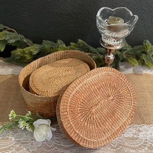 May include: Oval-shaped woven baskets with lids. The baskets are made of a light brown material and have a textured surface. One basket is open, revealing a smaller basket inside. The lid of the other basket is partially open. A candle holder and greenery are in the background.