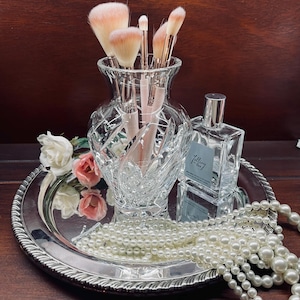 May include: A clear cut-glass vase containing five makeup brushes with light pink bristles, positioned next to a square glass perfume bottle labeled Falling in Love, a strand of white faux pearls, and two artificial roses on a silver tray.