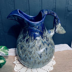 May include: A handcrafted ceramic pitcher with a unique, organic shape. The pitcher features a deep blue glaze with lighter, textured accents, creating a marbled effect. The handle is integrated into the design, and the pitcher sits on a white lace doily.