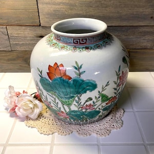 May include: A white ceramic ginger jar featuring hand-painted lotus flowers in shades of orange, pink, and green. The neck of the jar is decorated with a red and blue geometric border pattern. The jar has a rounded body and a wide opening.