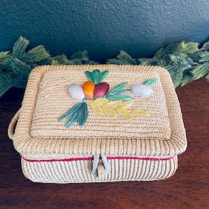 May include: A rectangular woven straw box with a lid, featuring a decorative floral design with colorful accents. The box has a red trim and a small handle. The lid is adorned with embroidered vegetables and flowers.