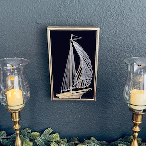 May include: A framed sailboat string art piece with a gold frame, set against a dark background. The sailboat is crafted with white and gold thread, depicting the sails and hull. Two glass candle holders with lit candles flank the artwork.