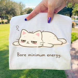 Puede incluir: Bolso tote blanco con un gato de dibujos animados con expresión de mal humor. El gato está tumbado. El texto "Bare minimum energy" está impreso en marrón debajo.