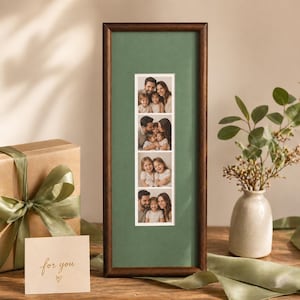 May include: A dark brown framed photo display with four family portraits against a green background. A gift box with a green ribbon and a card that says "for you" are in the foreground. A small vase with greenery is on the right.
