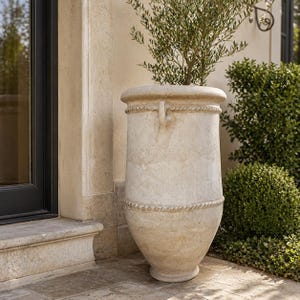 May include: A tall, beige outdoor planter with a rope-like design around the top and middle. The planter has a wide top and a narrower base, and it contains a small green plant. The planter is placed on a stone patio. The planter is made of stone.