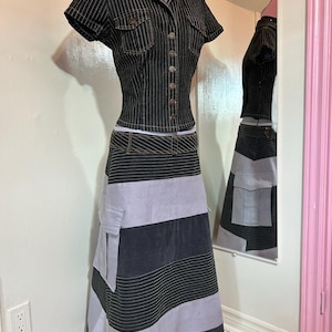 Peut inclure: Ensemble deux pièces sur mannequin. Le haut est une chemise boutonnée à manches courtes avec des fines rayures verticales et deux poches poitrine. La jupe est longue, à rayures, avec une poche sur le côté. La palette de couleurs comprend noir, gris et gris clair.