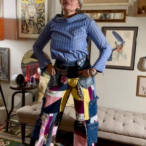 Può includere: Una persona indossa una camicia a righe blu e bianca e pantaloni in denim patchwork. I pantaloni presentano vari colori e motivi, tra cui denim, giallo, rosa e viola. Lo sfondo include opere d'arte incorniciate e un tappeto fantasia.