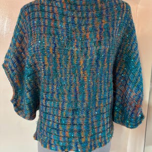 May include: A teal, orange, and blue woven top with sequin detailing. The top features a boat neck and three-quarter sleeves. The fabric has a textured look and is displayed on a dressmaker's dummy.