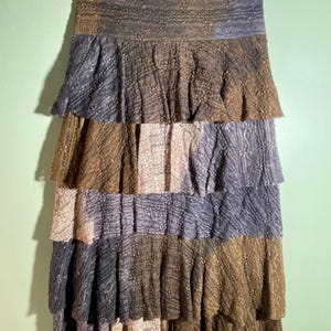 May include: A tiered skirt in shades of brown, grey, and beige. The skirt is made of a textured fabric and hangs from a black hanger. The skirt has a layered design with each tier slightly ruffled, creating a flowing effect.