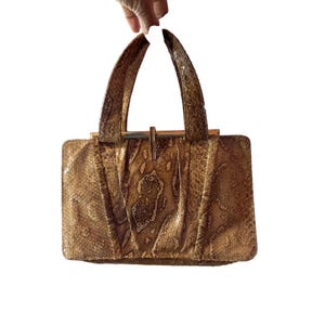 May include: A vintage handbag with a snakeskin pattern in shades of brown and tan. The bag has a rectangular shape with a structured design, featuring a gold-tone metal clasp and handles. The handles are arched and attached to the bag with gold-tone hardware.